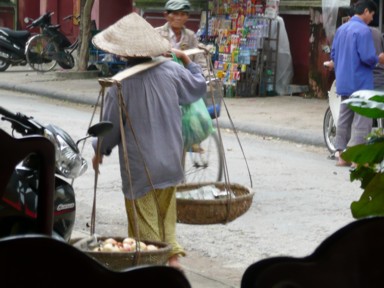 三角の帽子をかぶり籠を肩にかけて野菜を売る