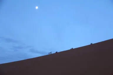 満月が照らす夕暮れの砂漠を登る人々